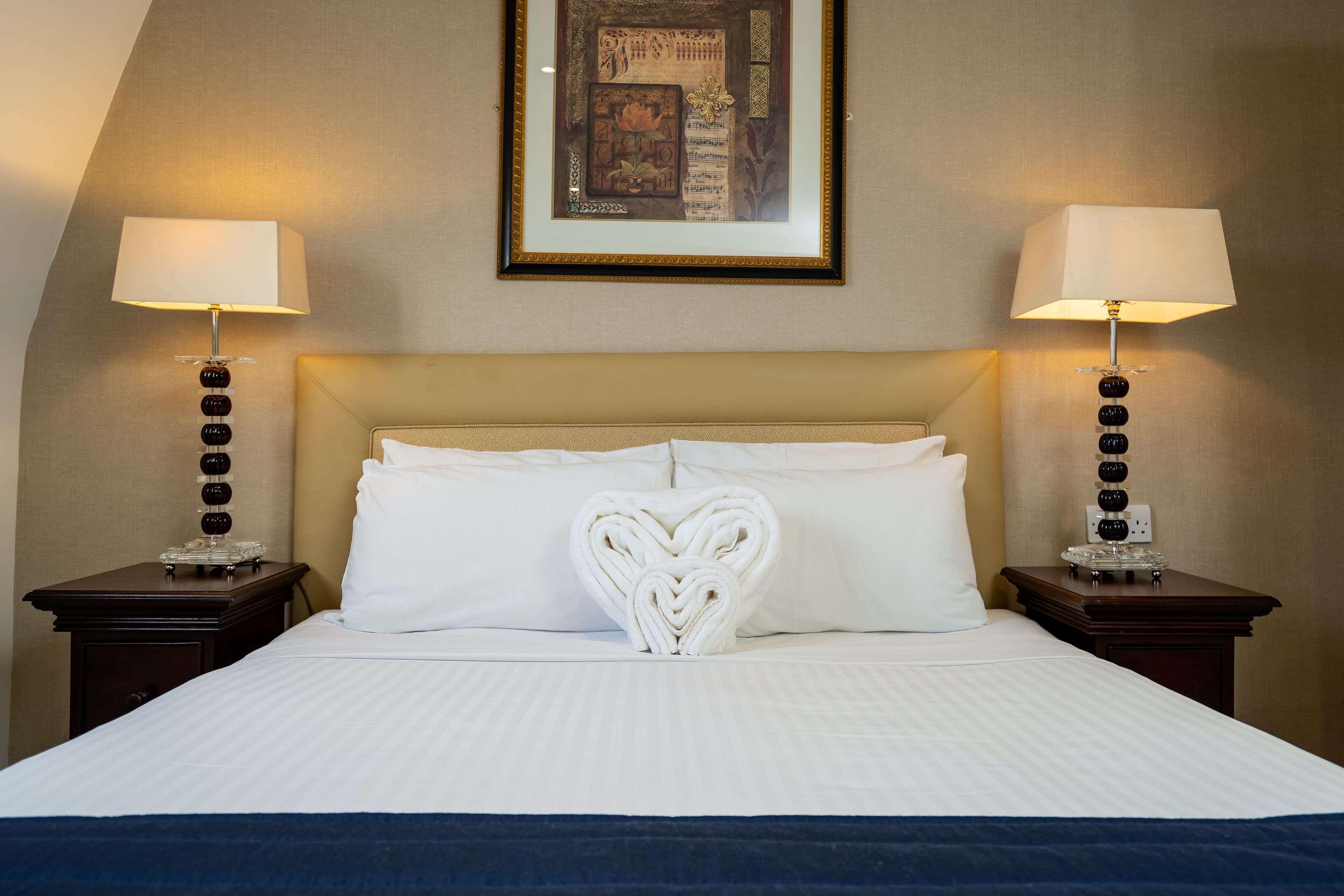 A hotel room with a neatly made bed, two bedside lamps, and a framed artwork on the wall.