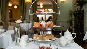 A three-tiered dessert stand with pastries and sandwiches, a white teapot, cups, and a vase with flowers on a table in a dining area. - The Clarendon Hotel - Blackheath (London)