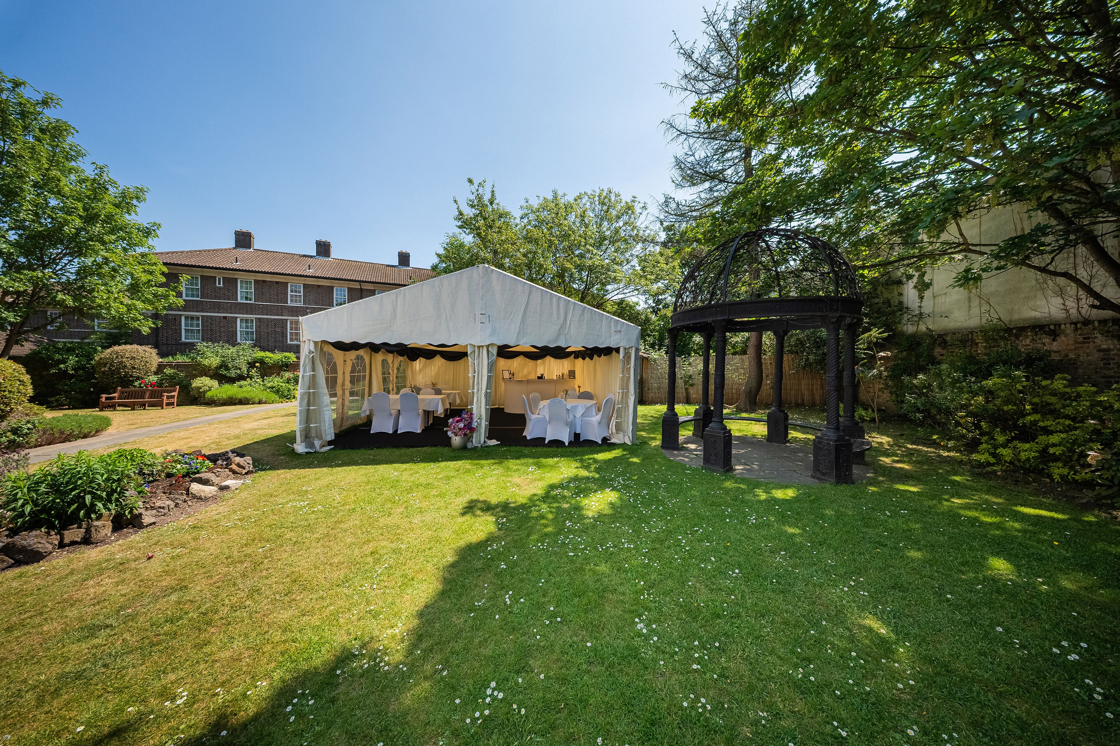 A tented outdoor area with tables and chairs, surrounded by a well-maintained lawn and trees.