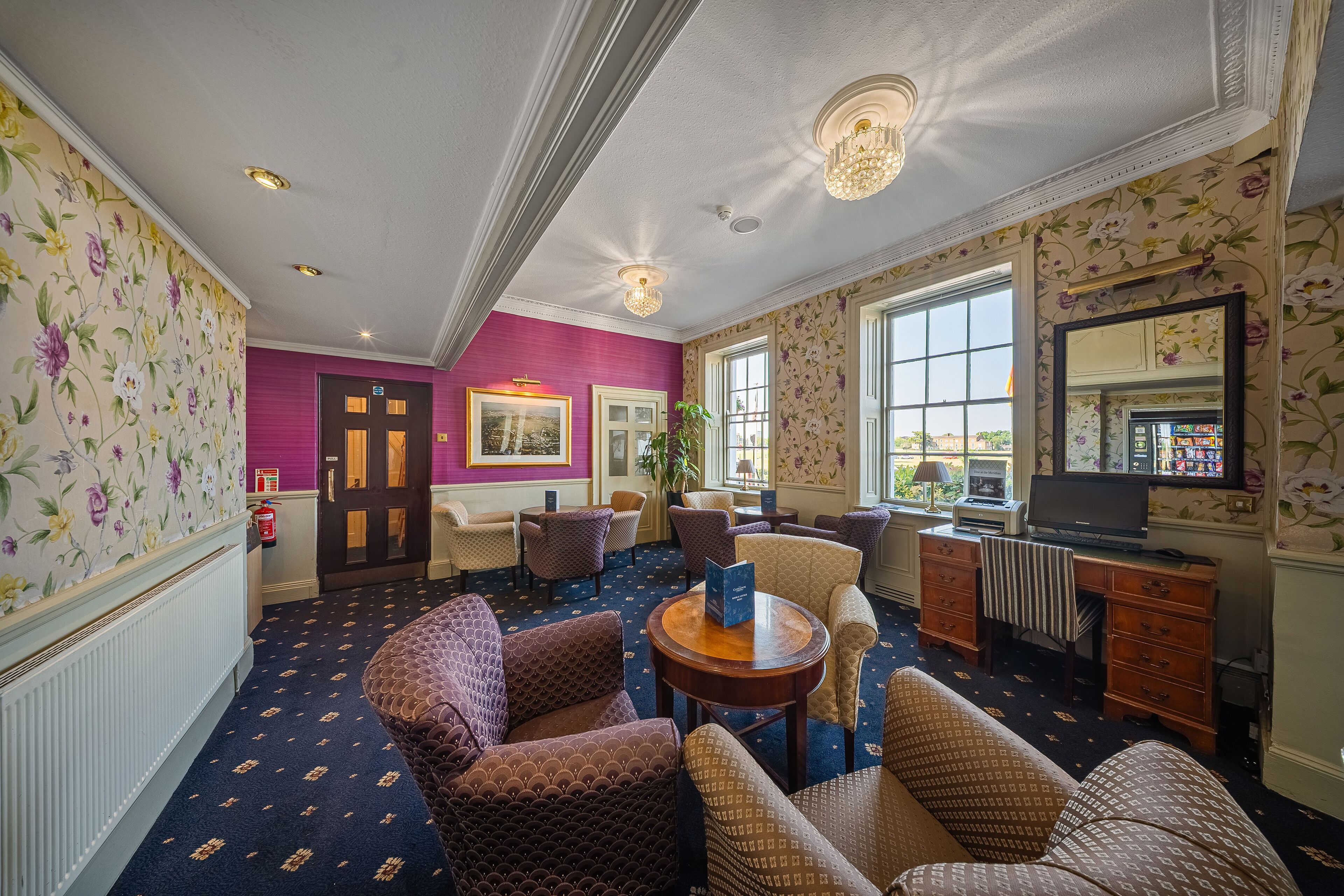 Una hall d'albergo con carta da parati decorata, una parete color viola e una zona relax con divani e poltrone a motivi.