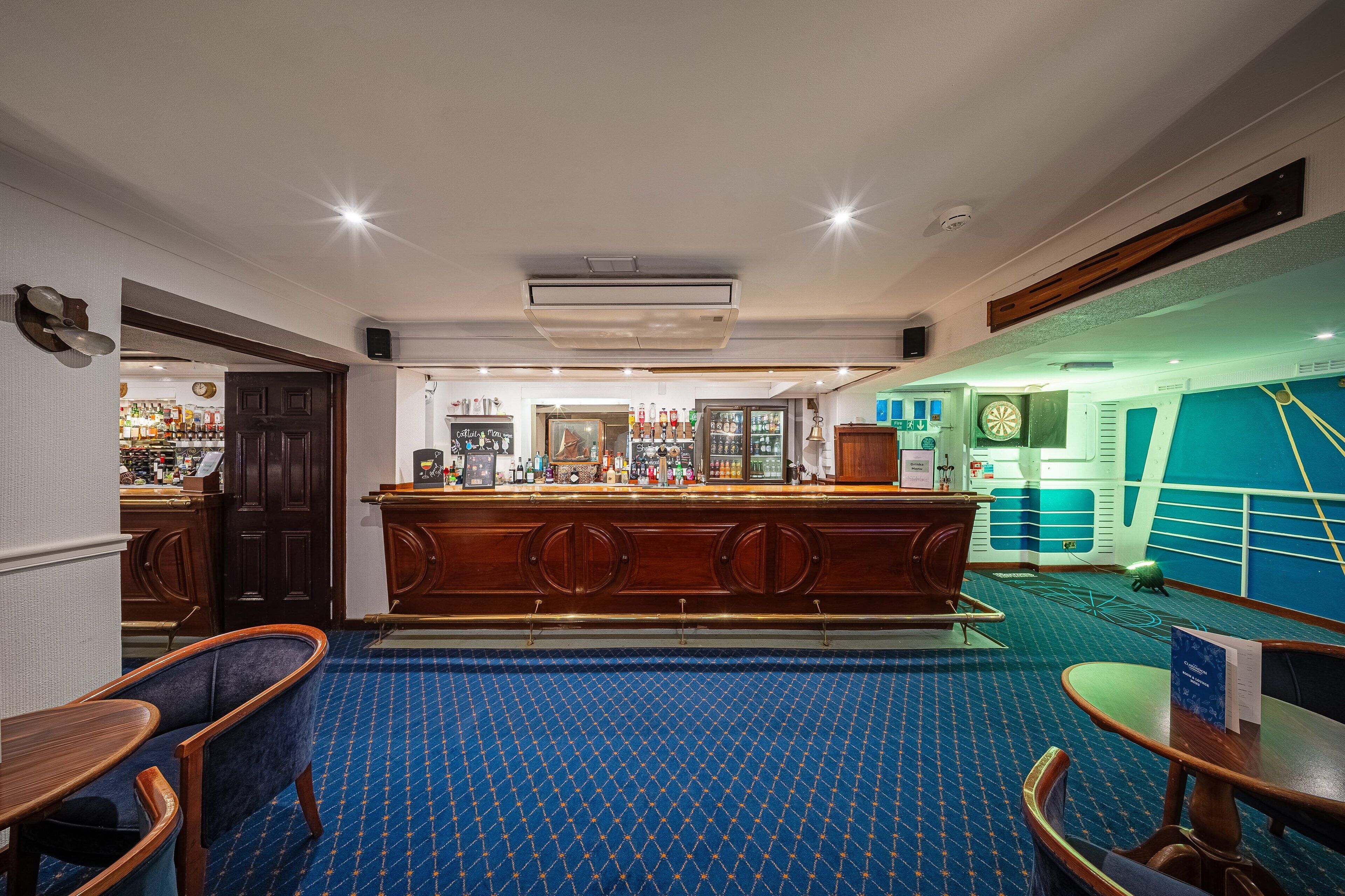 Zona de bar con alfombra estampada azul, mostrador de bar de madera y asientos con mesas y sillas de madera.