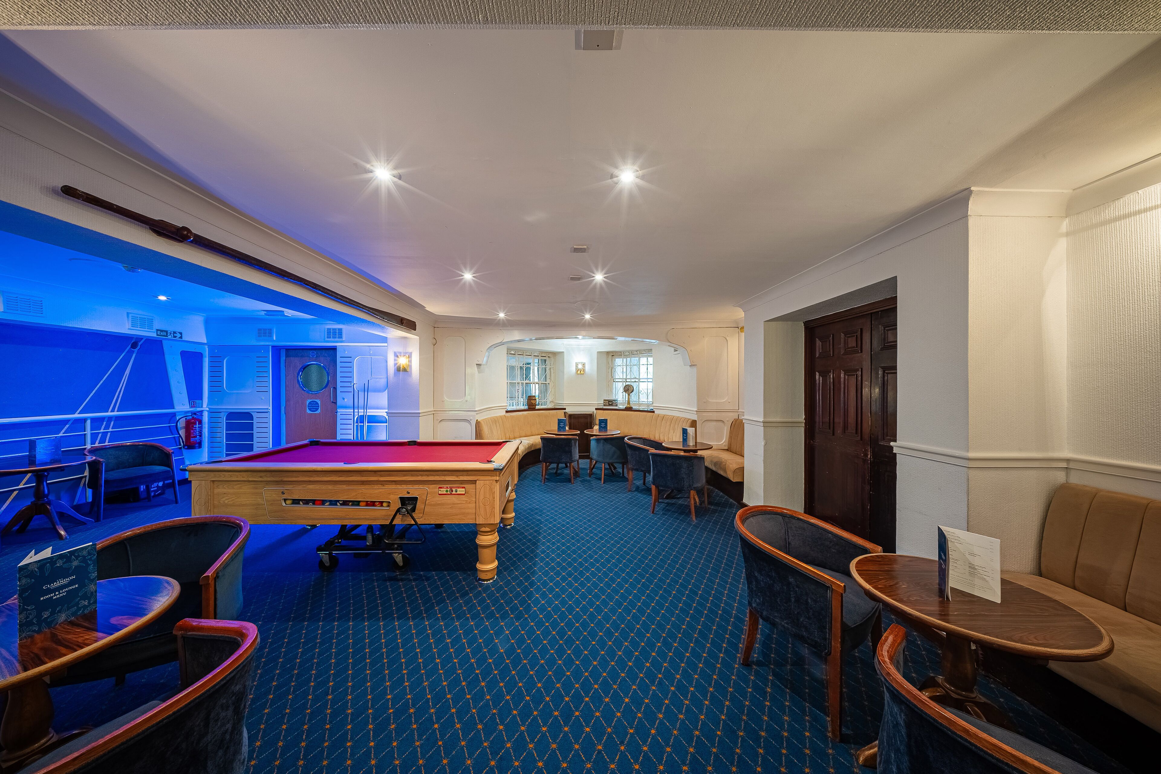 Une table de billard dans une pièce dotée d’une moquette bleue, de coins salon et d’une porte.