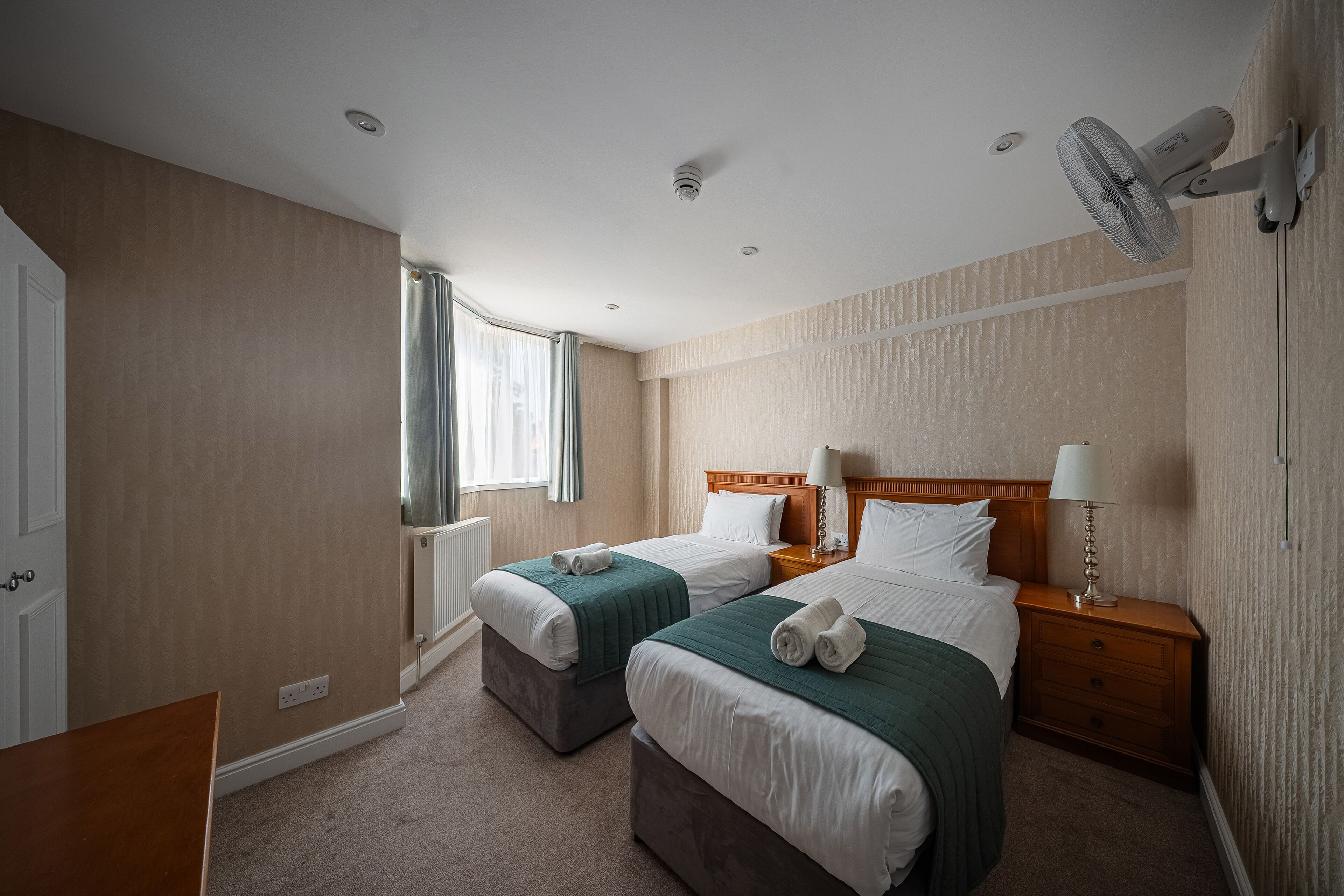 A hotel room with two beds, a desk, a chair, a window with curtains, a ceiling fan, and a nightstand with a lamp.