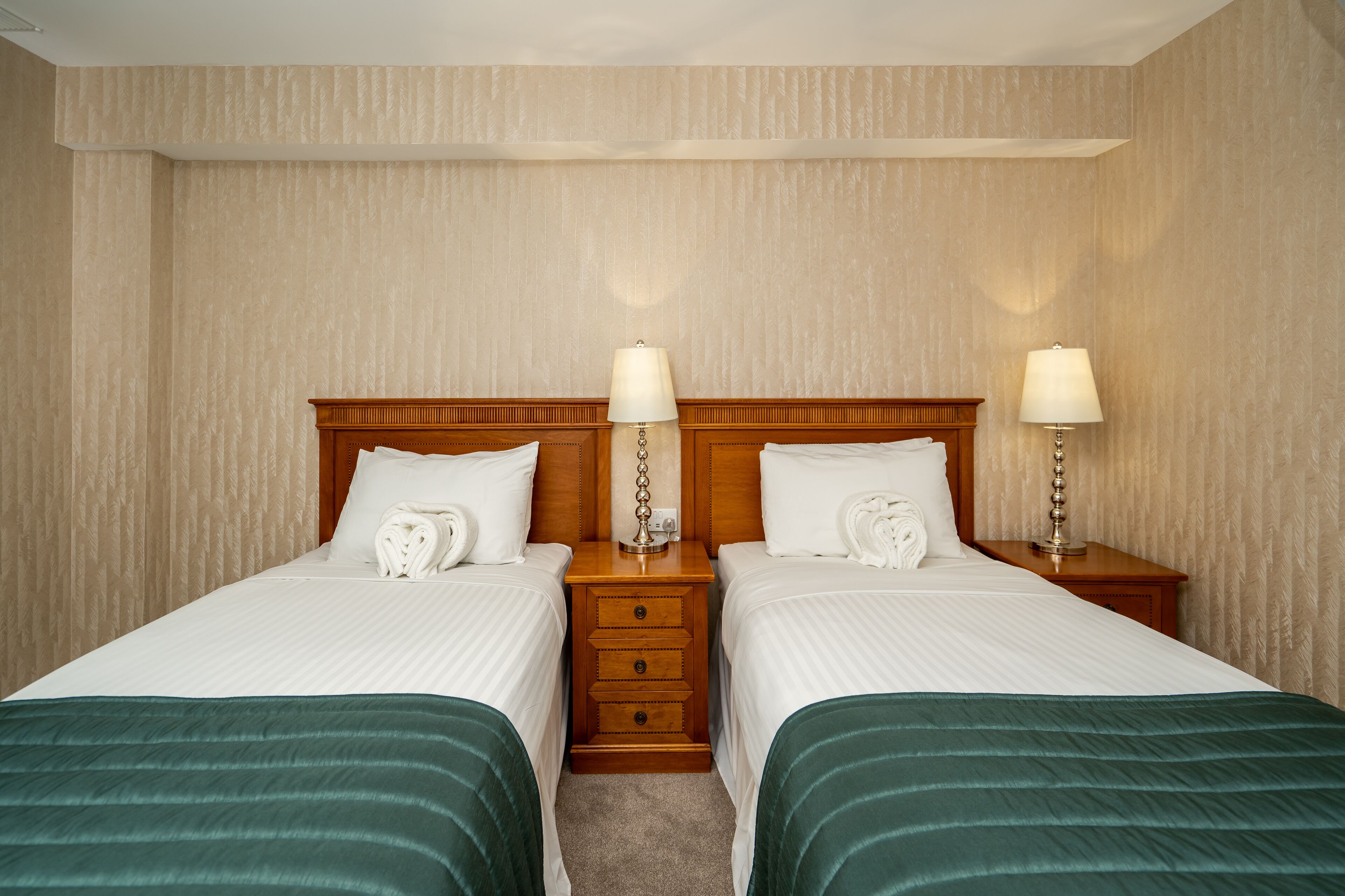A hotel room with two single beds, each with a wooden headboard, white linens, and green bedspreads. There are two bedside tables with lamps and a small drawer.