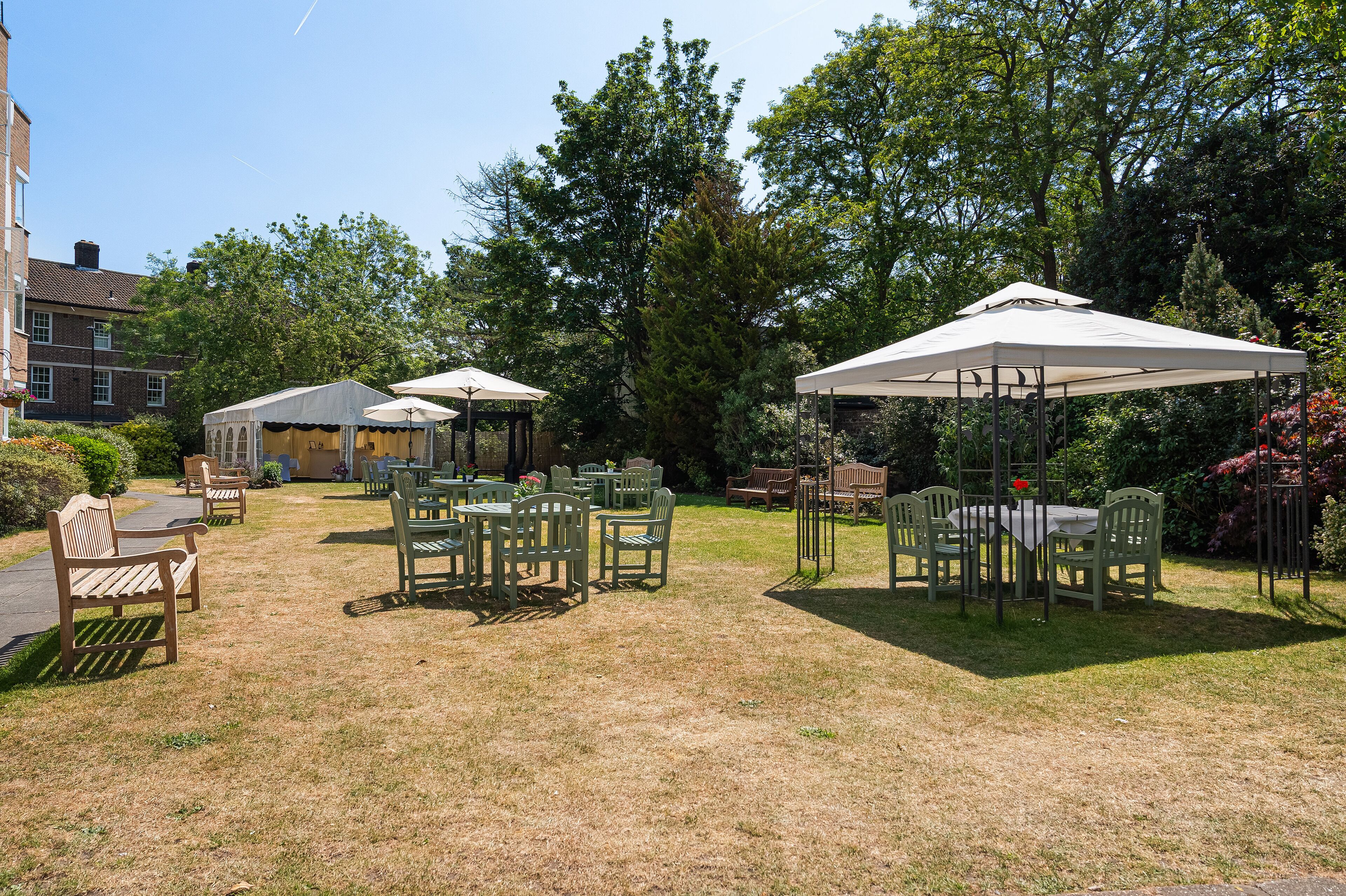 A garden patio area with tables and chairs under umbrellas, surrounded by trees and a brick building.
