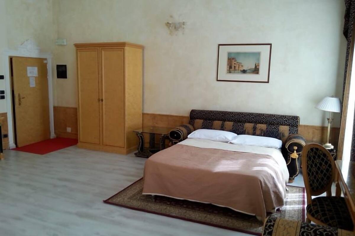A bedroom with a bed, a wardrobe, a small table, a chair, and a painting on the wall.