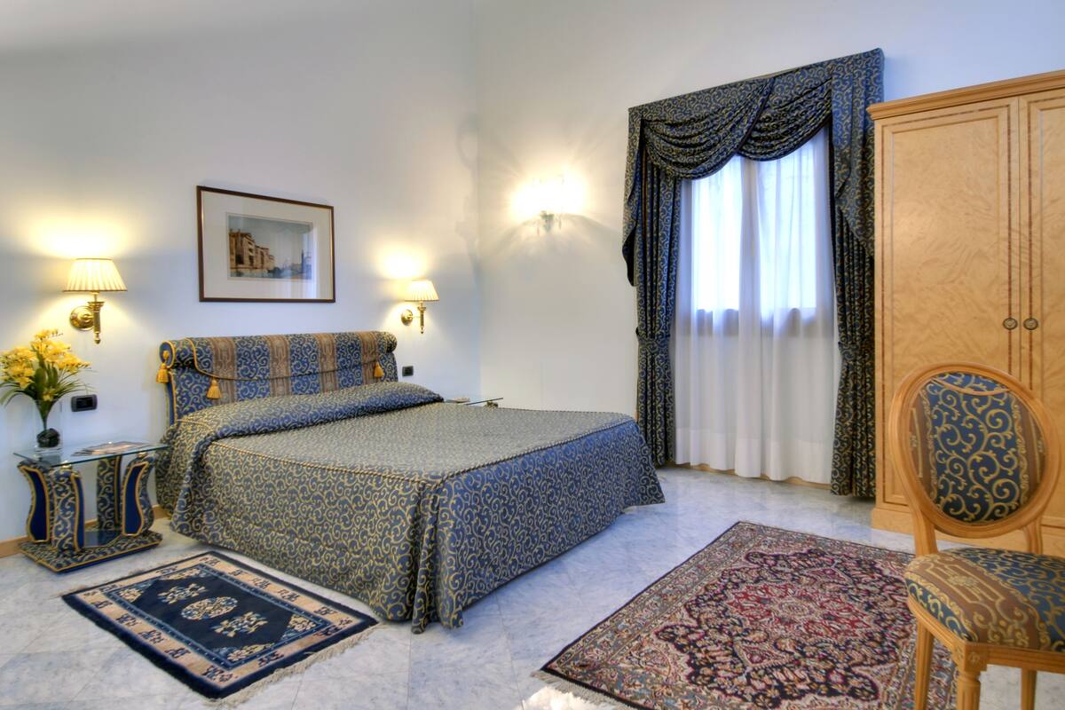 A bedroom with a large bed, two bedside lamps, a framed picture on the wall, a wooden wardrobe, and a chair.