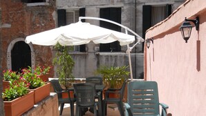 Un patio al aire libre con una mesa y sillas, un paraguas blanco, plantas en macetas y una pared de ladrillo.