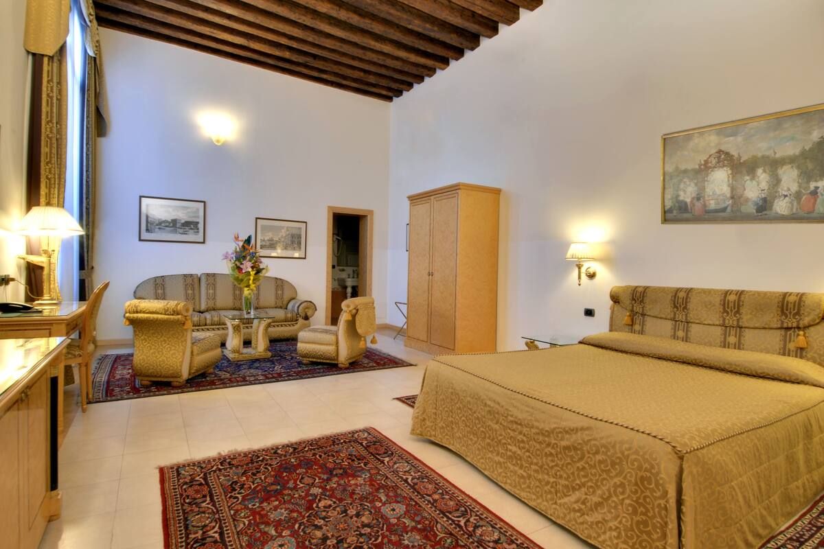A spacious bedroom with a large bed, a sofa, a chair, and a cabinet. There are two framed pictures on the wall, a lamp, and a painting.
