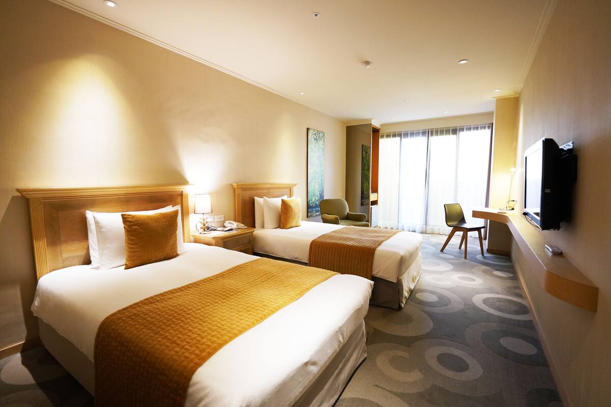 A hotel room with two beds, a desk, a chair, a television, and a large window.