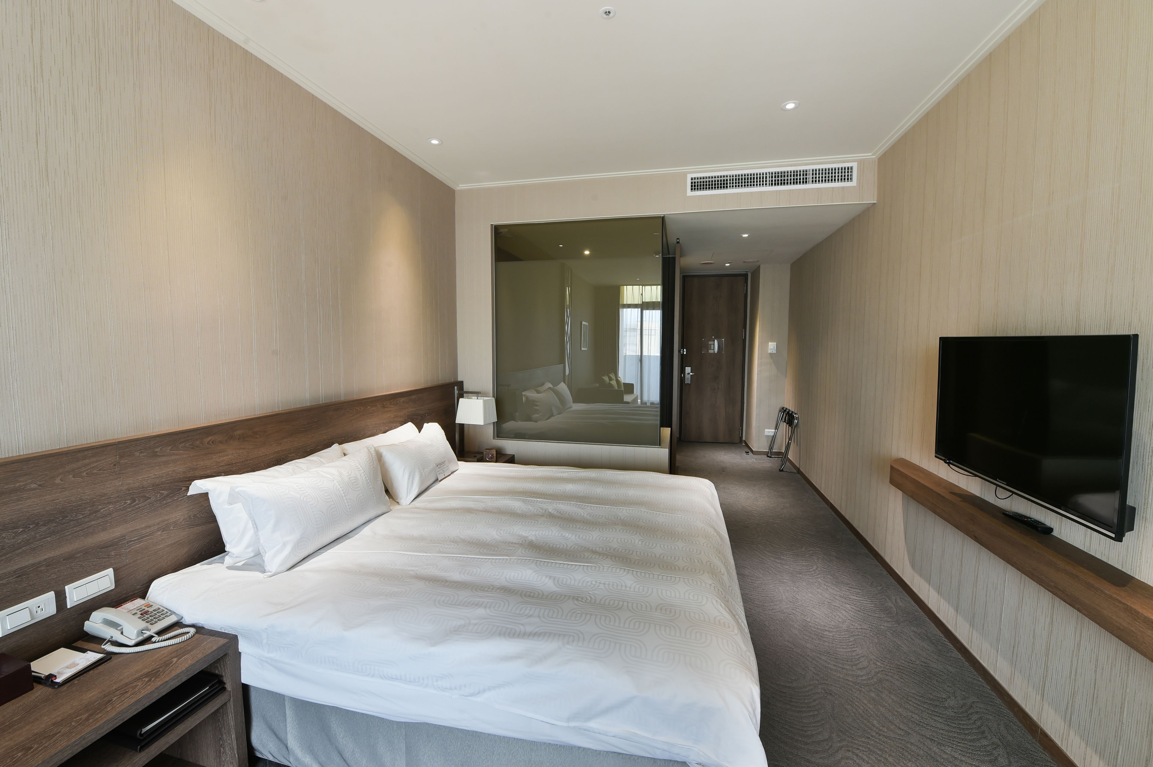 A hotel room with a large bed, a bedside telephone, a flat-screen TV mounted on the wall, and a door leading to a hallway.