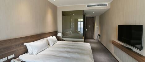 A hotel room with a large bed, a bedside telephone, a flat-screen TV mounted on the wall, and a door leading to a hallway.