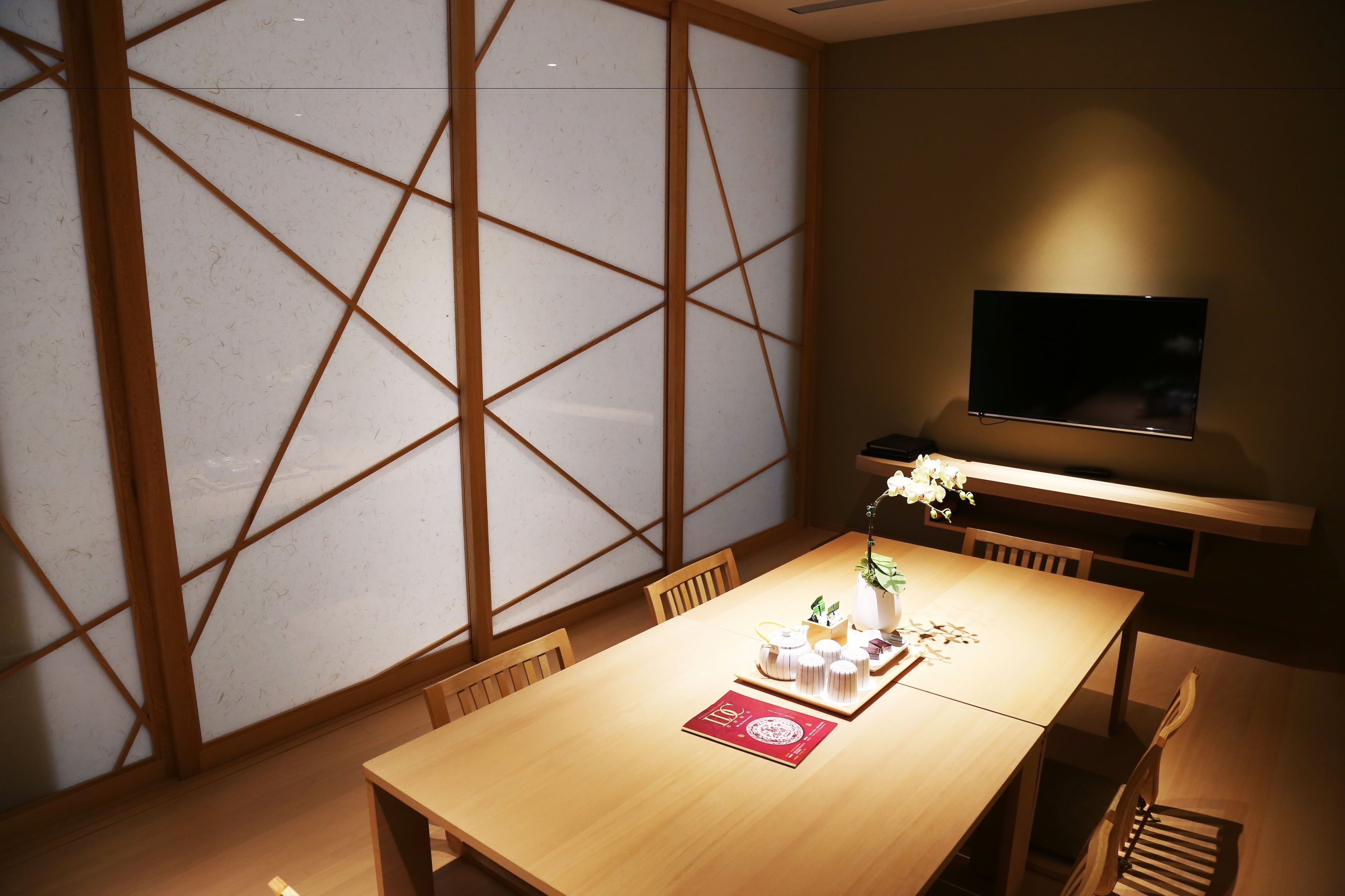 A dining area with a wooden table, chairs, a TV mounted on the wall, and a decorative vase with flowers.