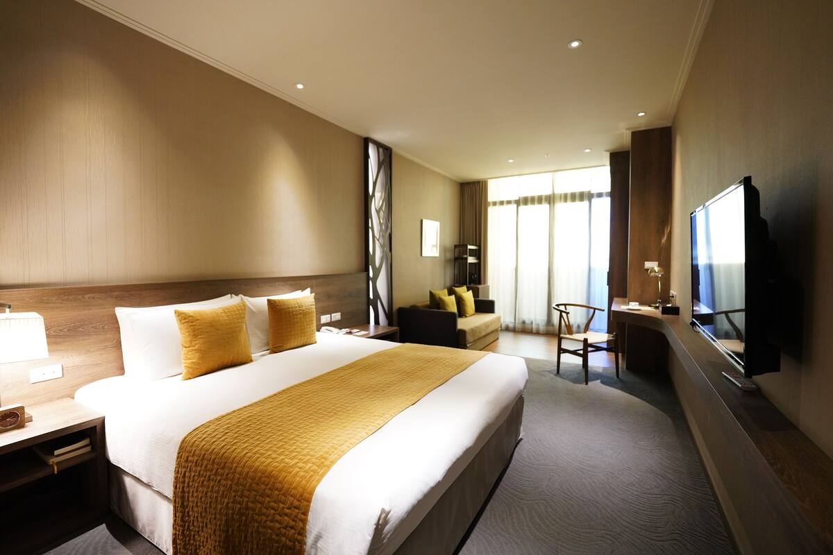 A hotel room with a large bed, a desk with a lamp, a TV, and a seating area with a sofa and chairs.