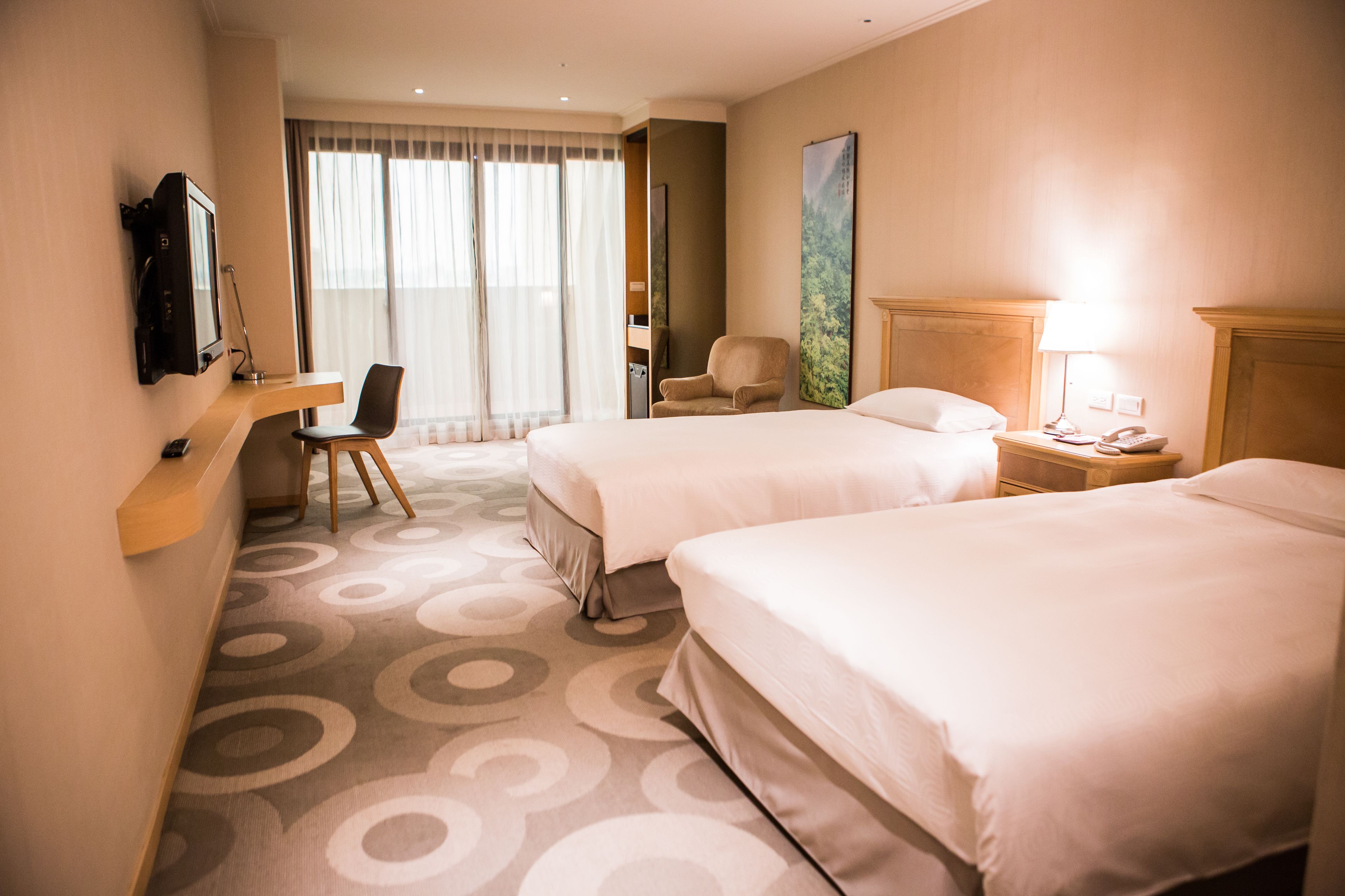 A hotel room with two beds, a desk, a chair, a TV, and a lamp.