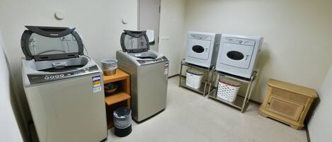 A laundry room with two washing machines, a dryer, a trash bin, and a wooden cabinet.