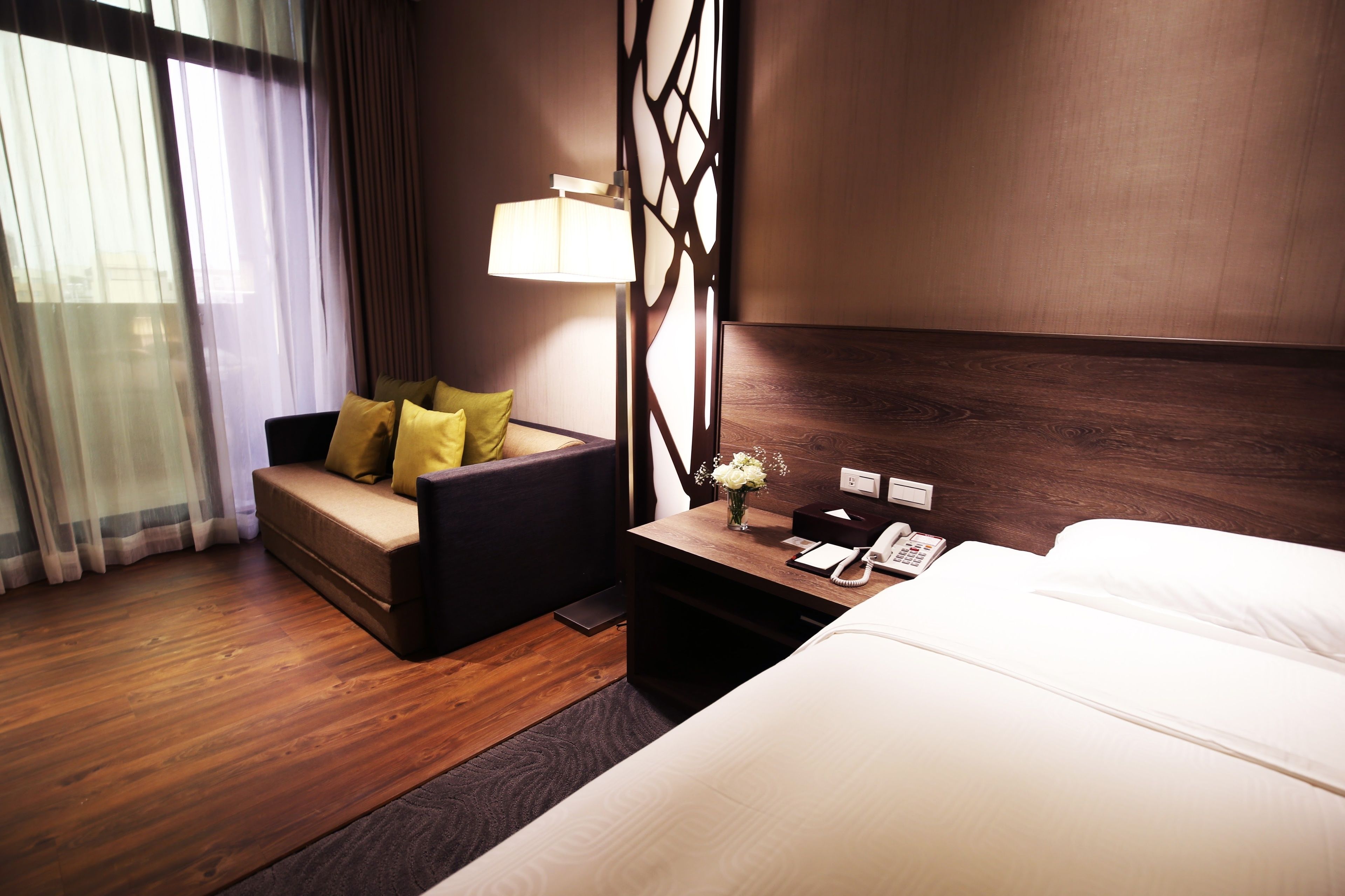 A hotel room with a bed, a sofa with yellow cushions, a nightstand with a lamp, and a wooden headboard.
