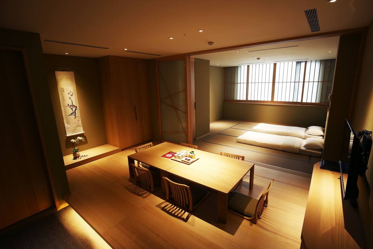 A modern hotel room with a wooden dining table, chairs, a sofa, and a large window with blinds.