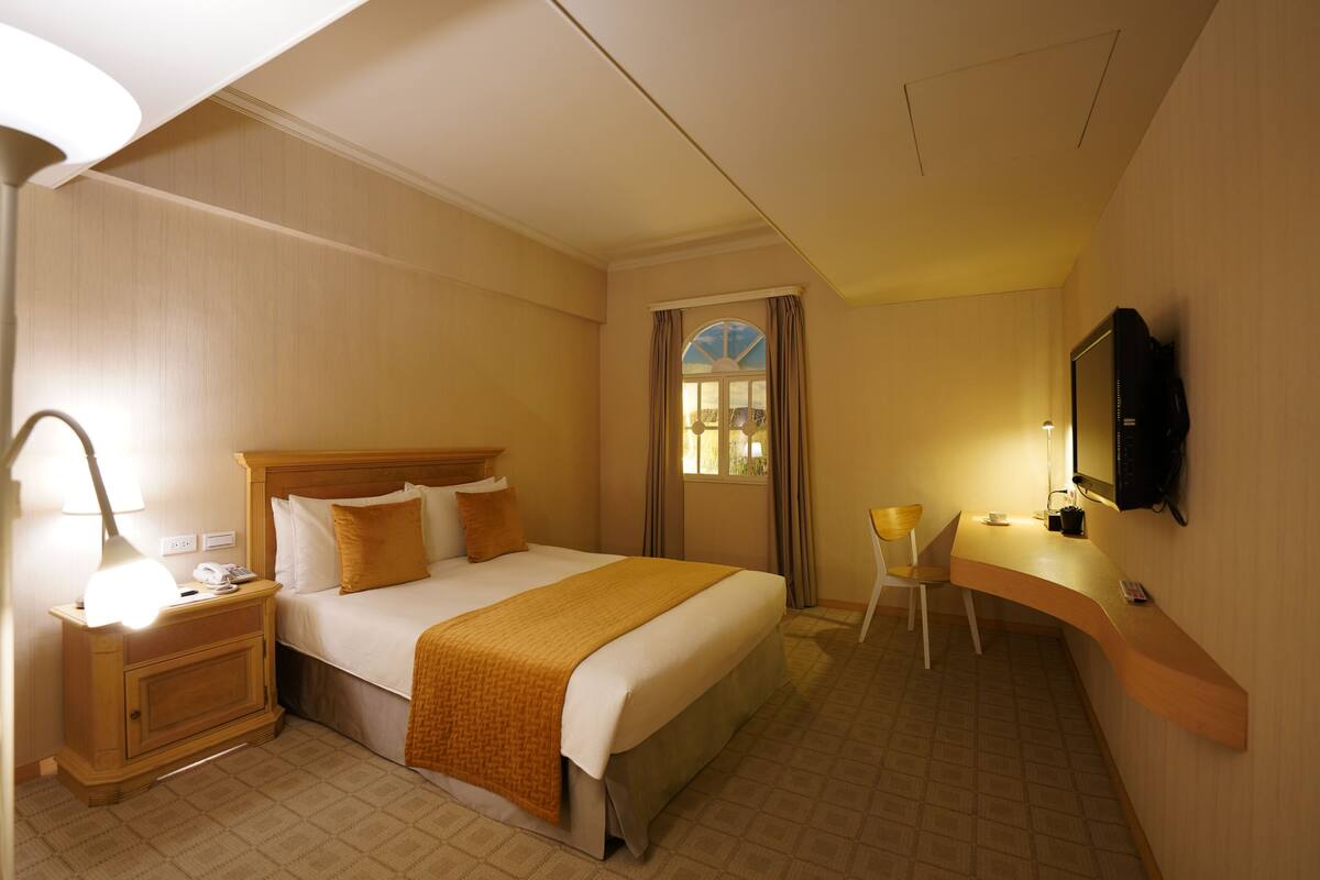 A hotel room with a bed, bedside table, lamp, chair, desk, and TV.