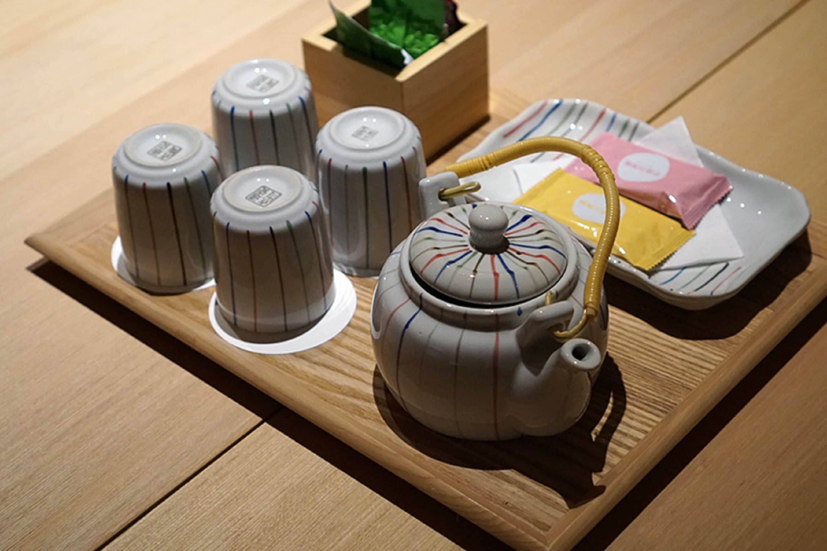 A wooden tray with a teapot, four cups, and a plate with packets.