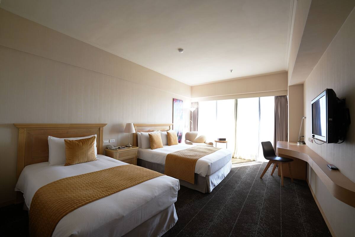 A hotel room with two beds, a desk, a chair, a television, and a large window with curtains.