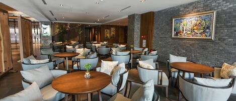 A modern hotel lobby with round tables and cushioned chairs, a large abstract painting on the wall, and a staircase leading to another level.