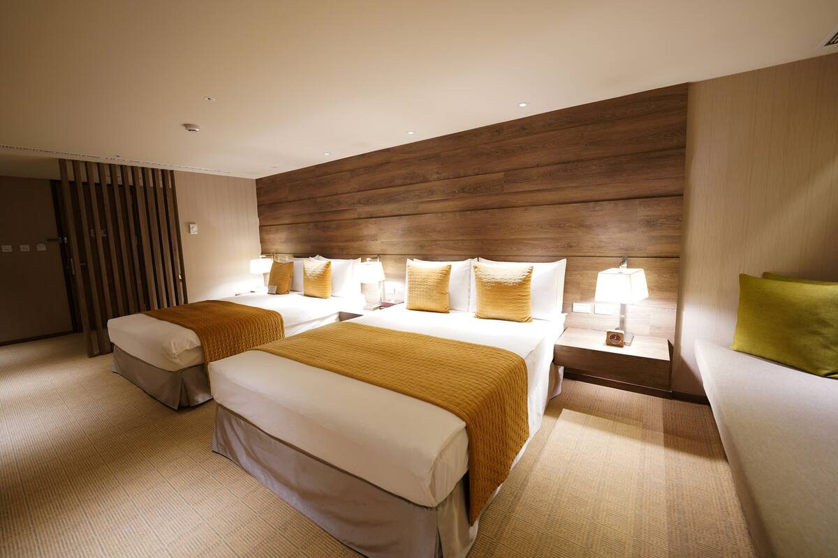 A hotel room with two beds, a sofa, a wooden headboard, and a nightstand with a lamp.
