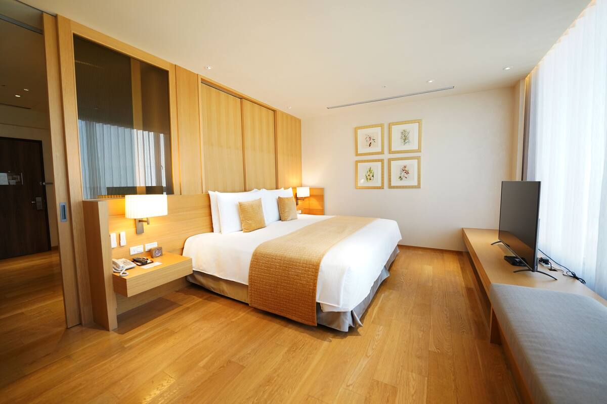 A modern hotel room with a large bed, wooden flooring, a bedside table with a lamp, a TV, and framed pictures on the wall.