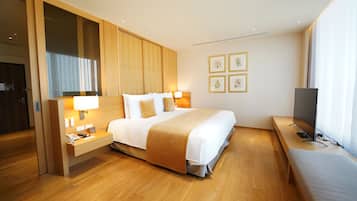 A modern hotel room with a large bed, wooden flooring, a bedside table with a lamp, a TV, and framed pictures on the wall.