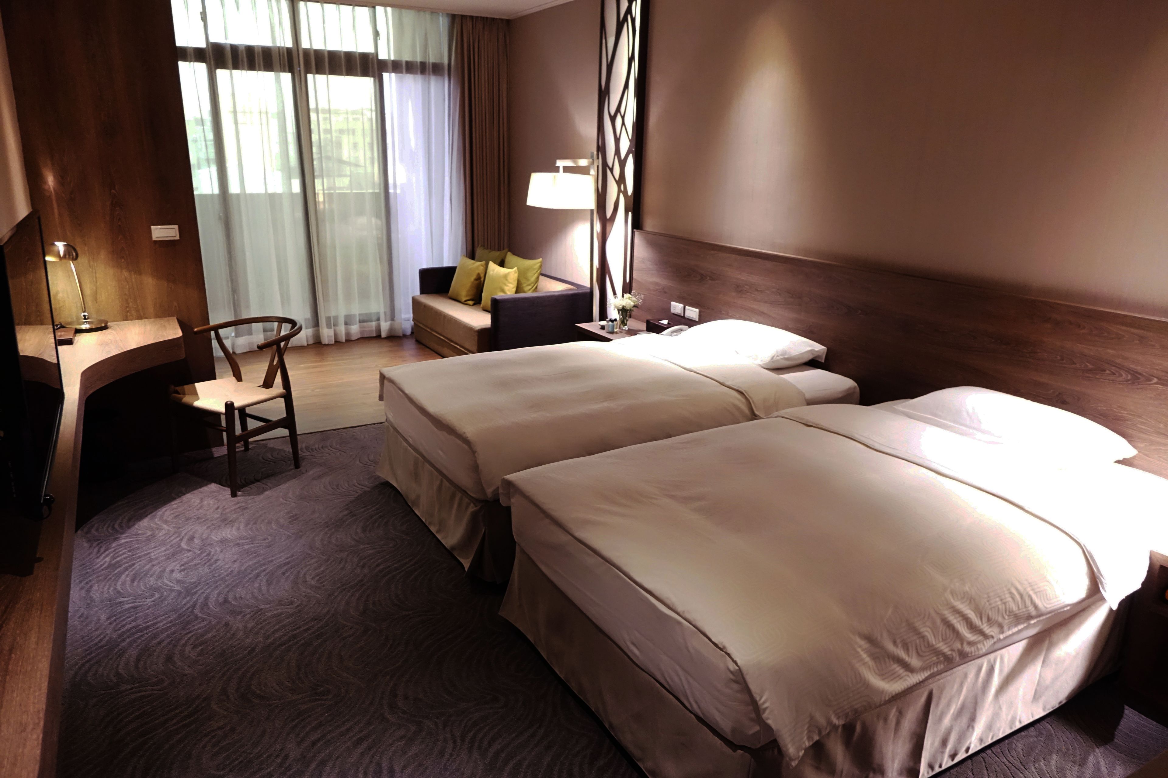 Quarto de hotel com duas camas, uma escrivaninha com cadeira, um sofá com almofadas, uma luminária e uma janela grande com cortinas.
