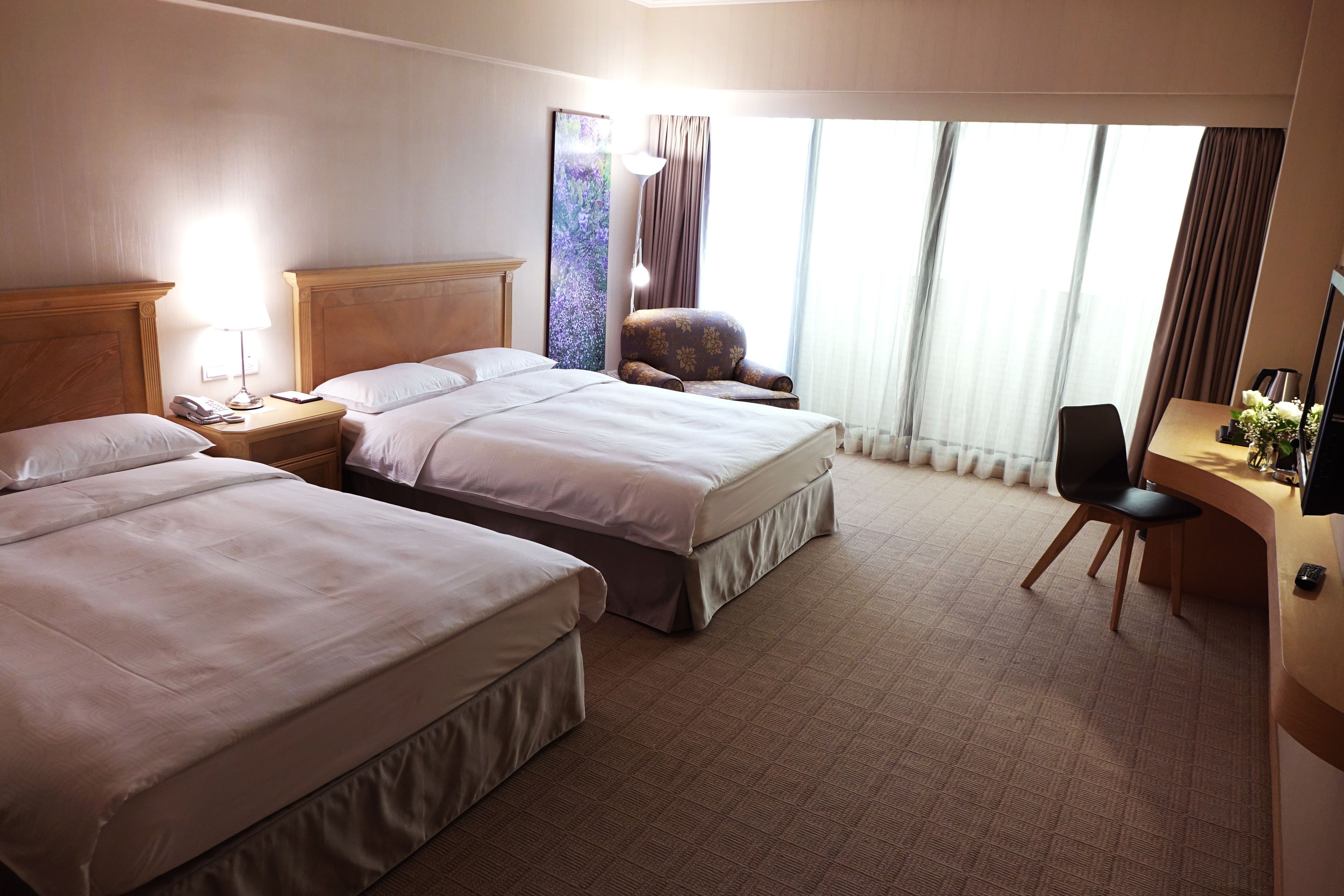 A hotel room with two beds, a desk, a chair, a lamp, and a large window with curtains.