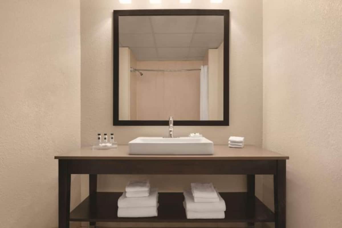 A bathroom with a rectangular sink, a mirror above it, and a shelf below holding folded towels.