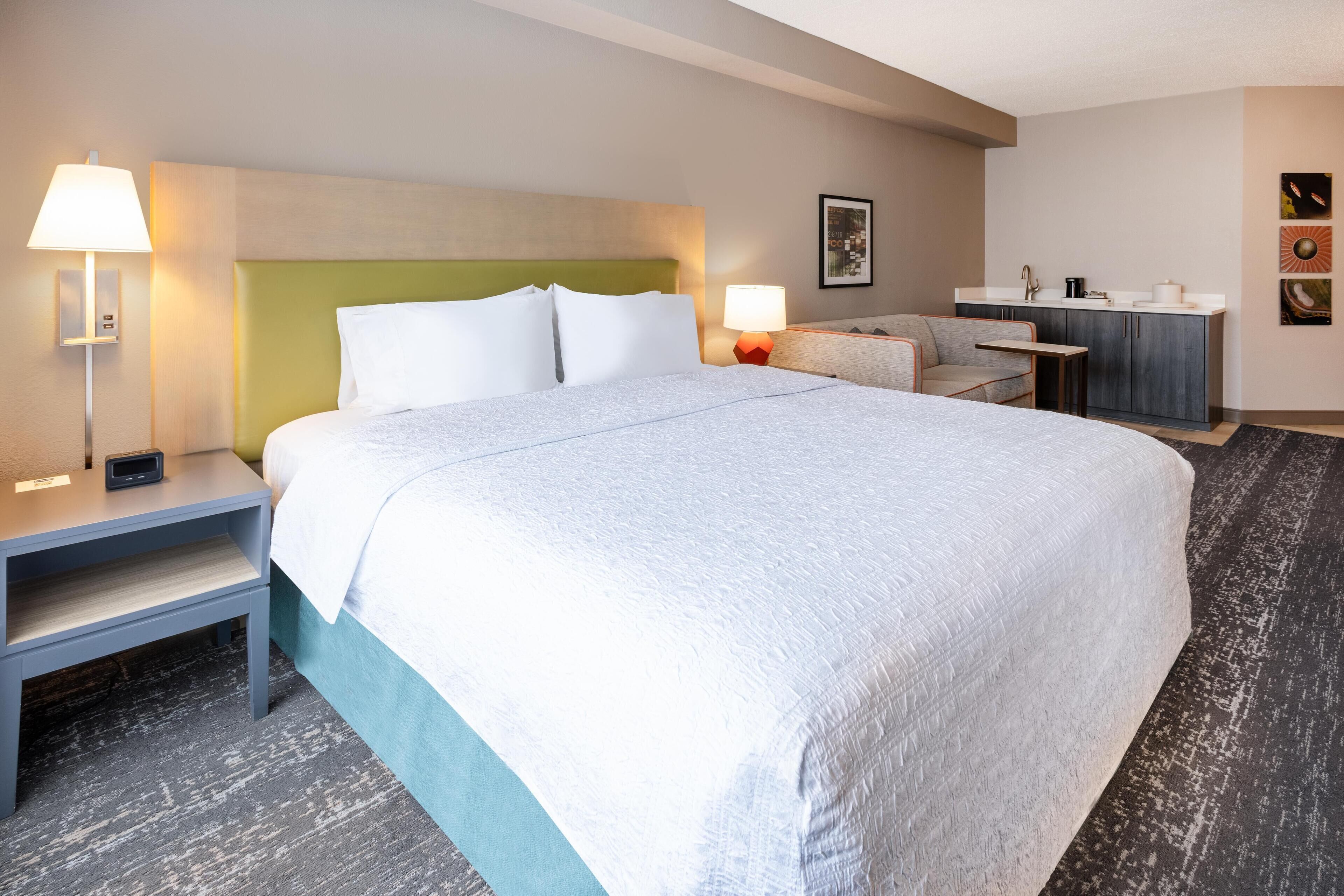 A hotel room with a large bed, bedside tables, a desk, and a kitchenette.