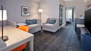 A hotel room with a sofa, armchair, and a desk with a lamp. There is a television and a door leading to the bathroom.