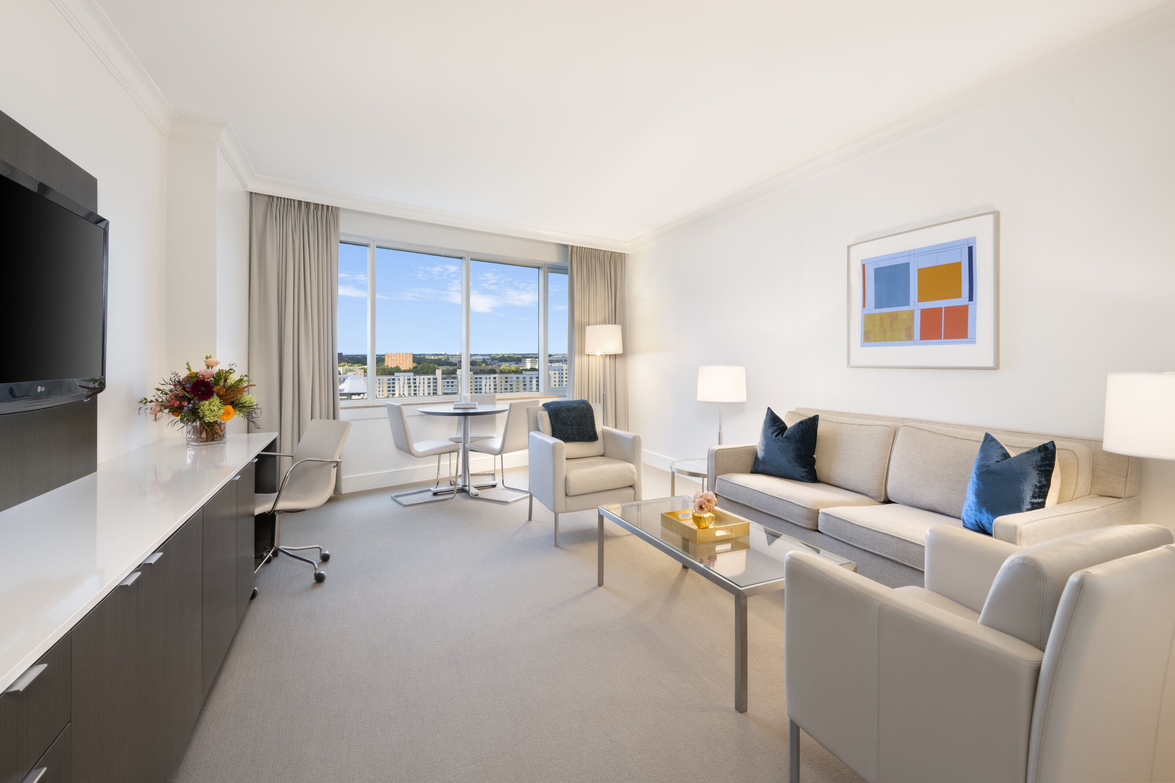 A modern hotel room with a flat-screen TV, a sofa, a coffee table, a dining table with chairs, and a large window offering a city view.