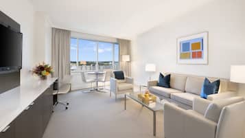 A modern hotel room with a flat-screen TV, a sofa, a coffee table, a dining table with chairs, and a large window offering a city view.