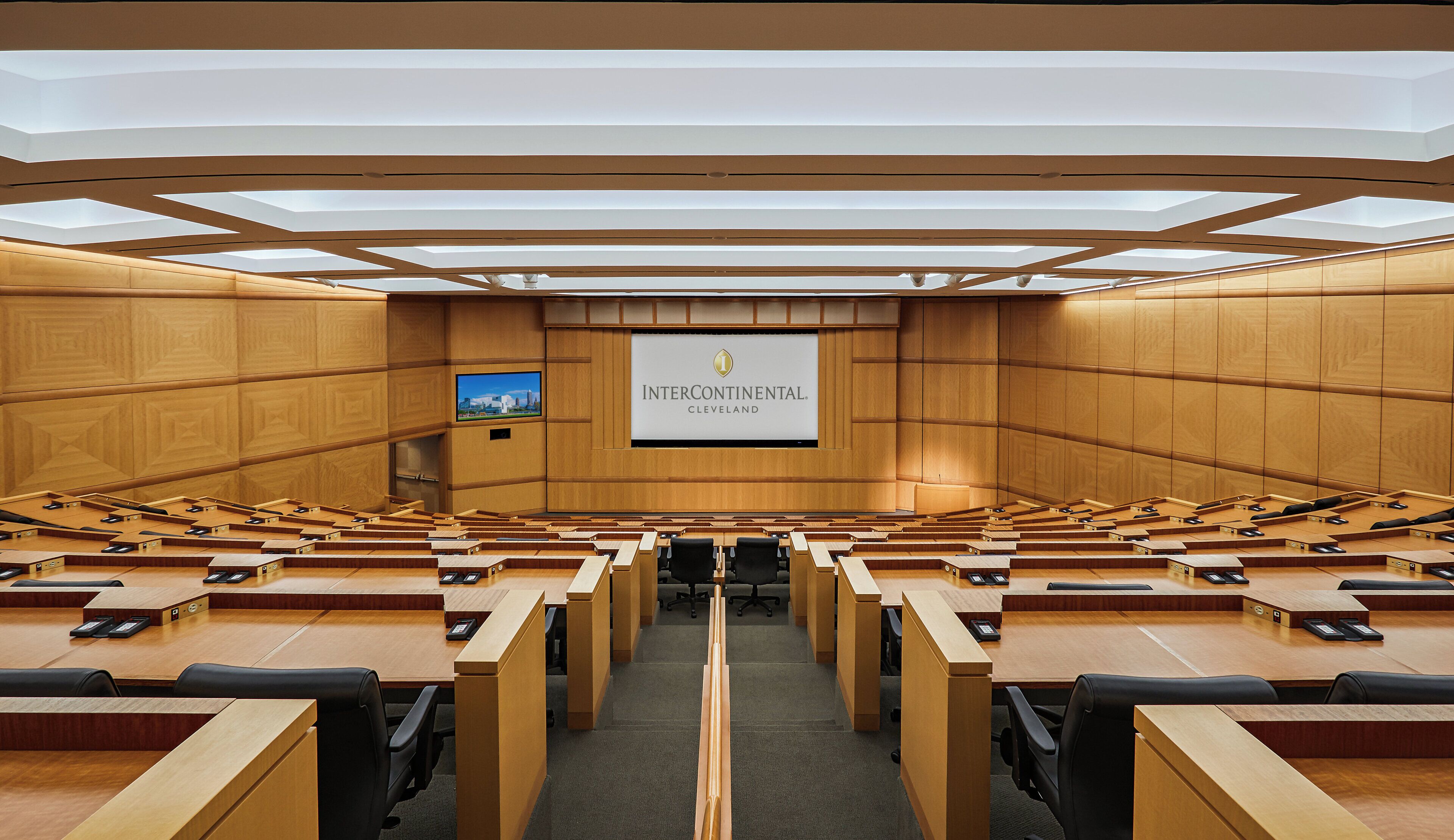 Un salón de conferencias con varias filas de escritorios y sillas, una pantalla grande mostrando un logotipo y un techo con tragaluces.