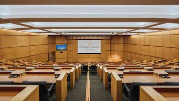 Un salón de conferencias con varias filas de escritorios y sillas, una pantalla grande mostrando un logotipo y un techo con tragaluces.