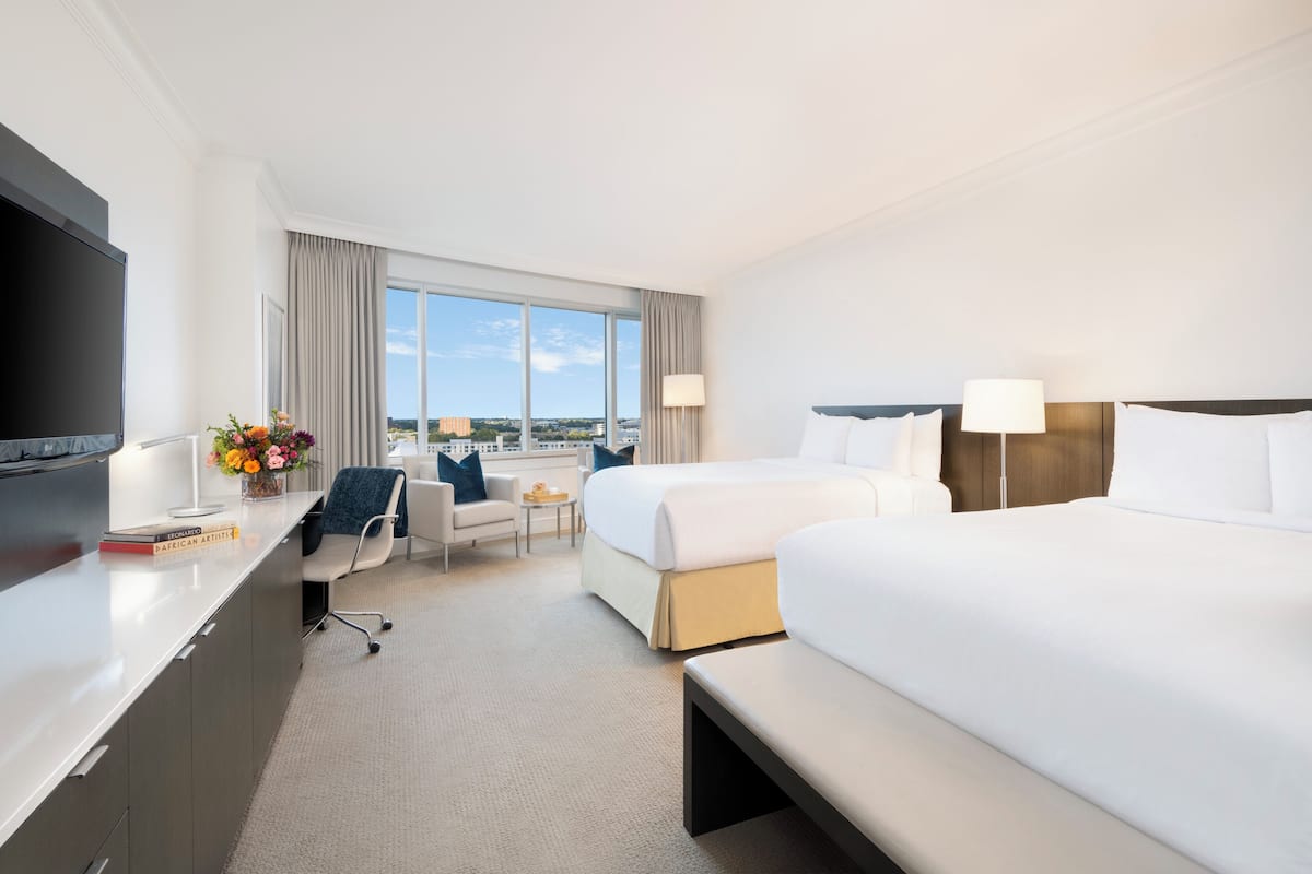 A hotel room with two beds, a desk with a TV, a chair, a small table with a vase of flowers, and a view of the city.