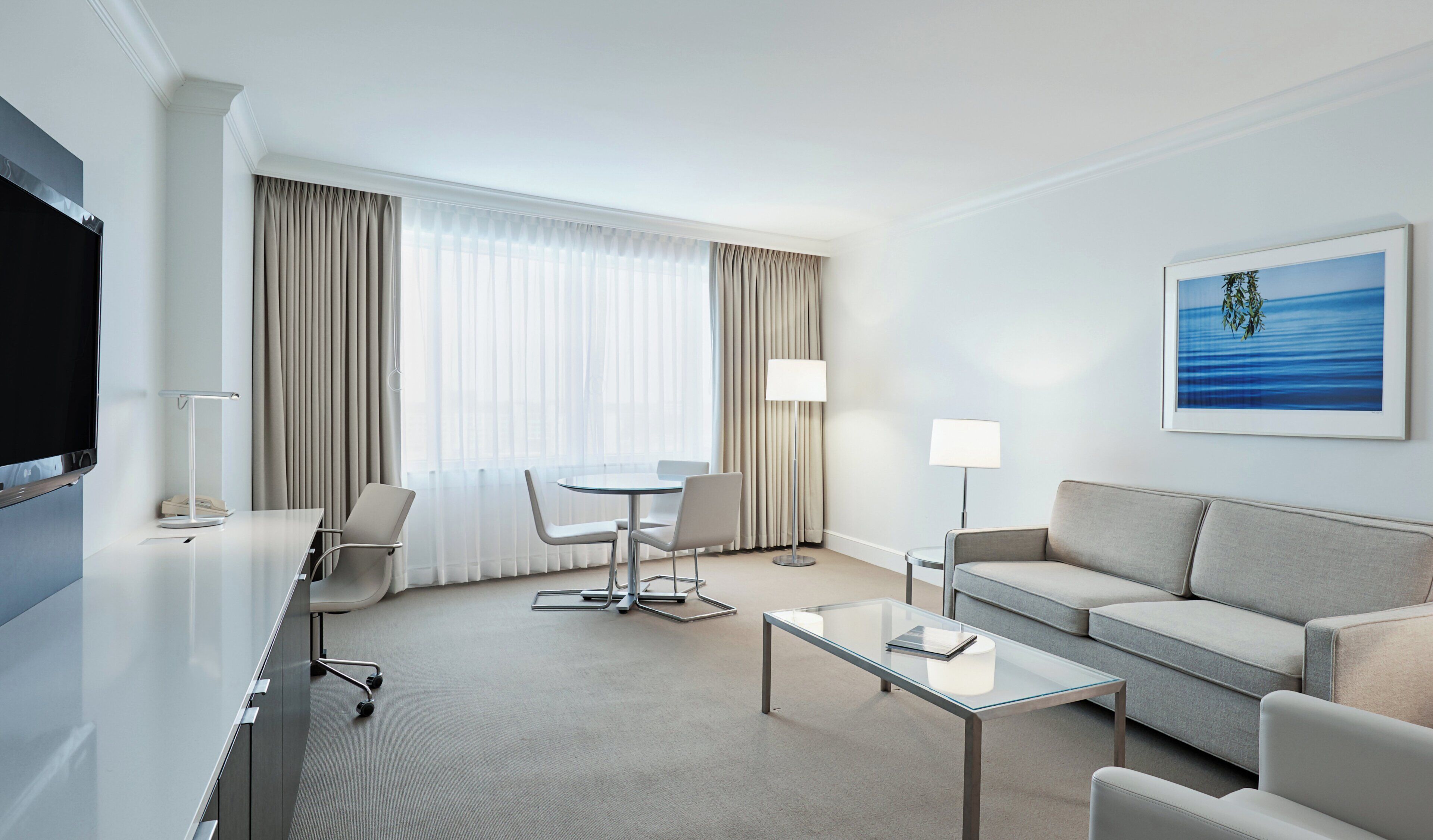 A hotel room with a flat-screen TV, a desk with a chair, a sofa, a coffee table, a dining table with chairs, and a large window with curtains.