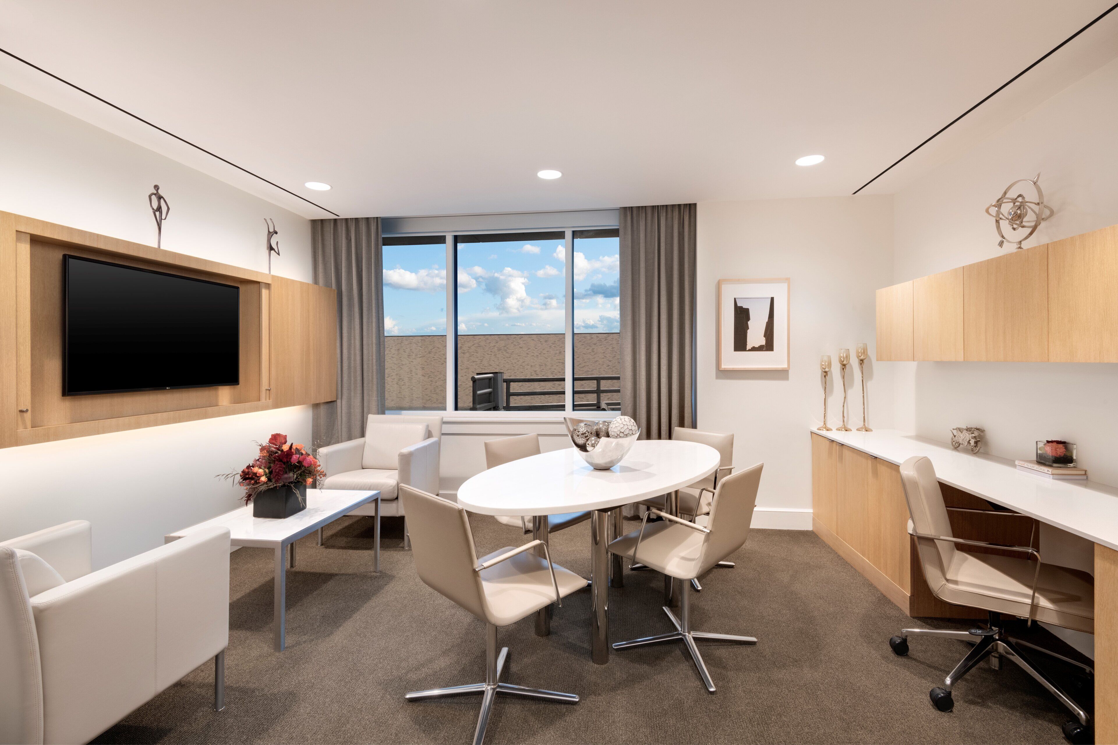 A modern conference room with a large table, white chairs, a flat-screen TV, and a large window with a view of the sky.