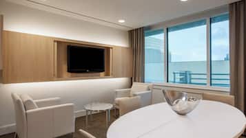 Una habitación de hotel moderna con televisor de pantalla plana montado en un panel de madera, una mesa redonda de comedor con un centro de mesa metálico, dos sillones blancos y una pequeña mesita auxiliar.