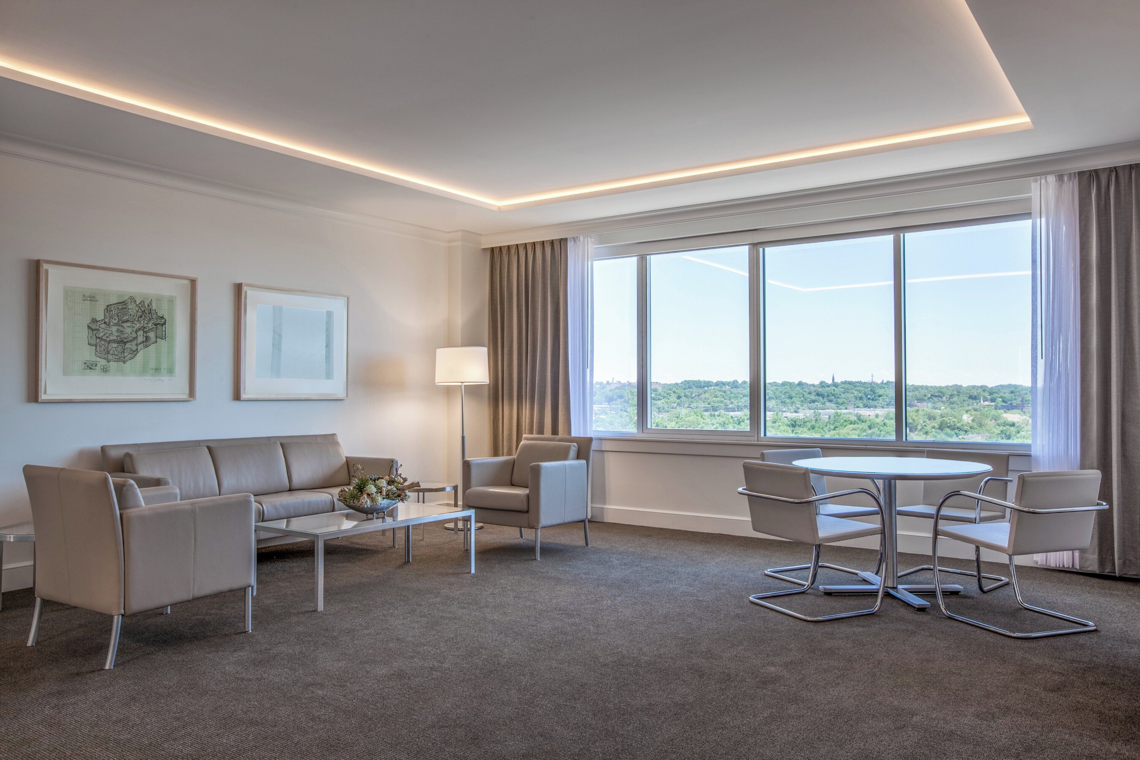Una habitación de hotel moderna con sofá, sillones, una mesa de centro y una mesa de comedor. Hay ventanas amplias con cortinas, un televisor de pantalla plana y cuadros en la pared.