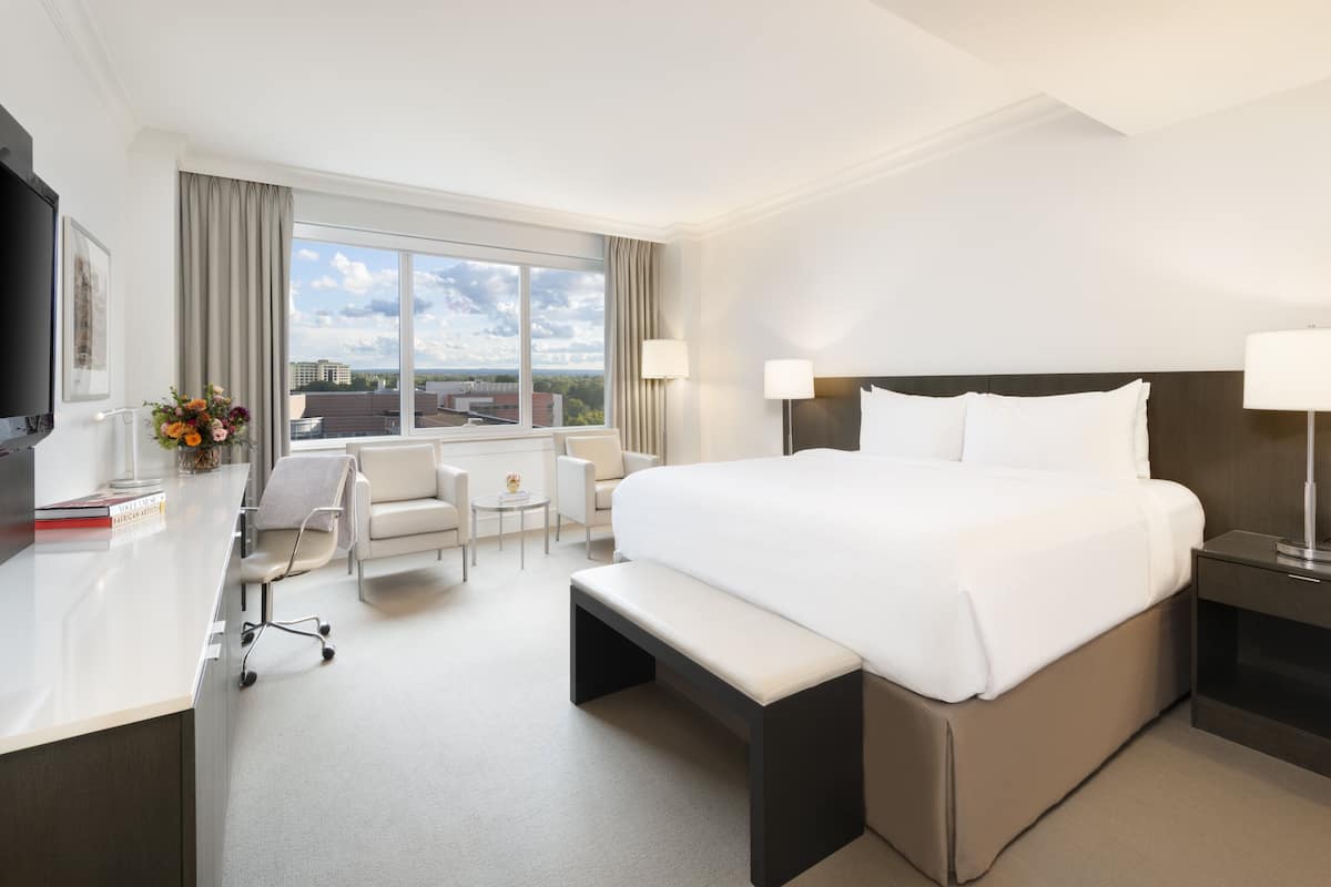 A hotel room with a large bed, a desk, a chair, a bench, and a TV.