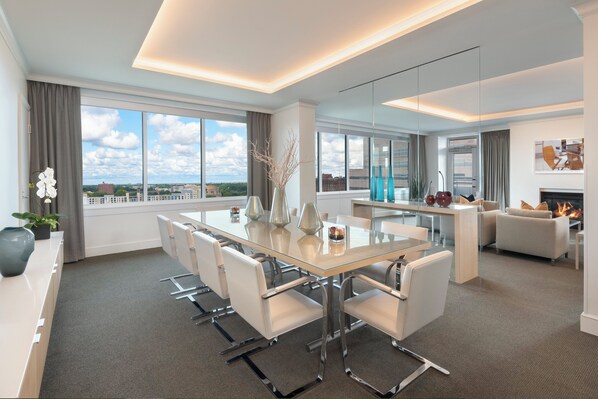 A modern hotel room with a glass dining table, white chairs, a sofa, and a fireplace.