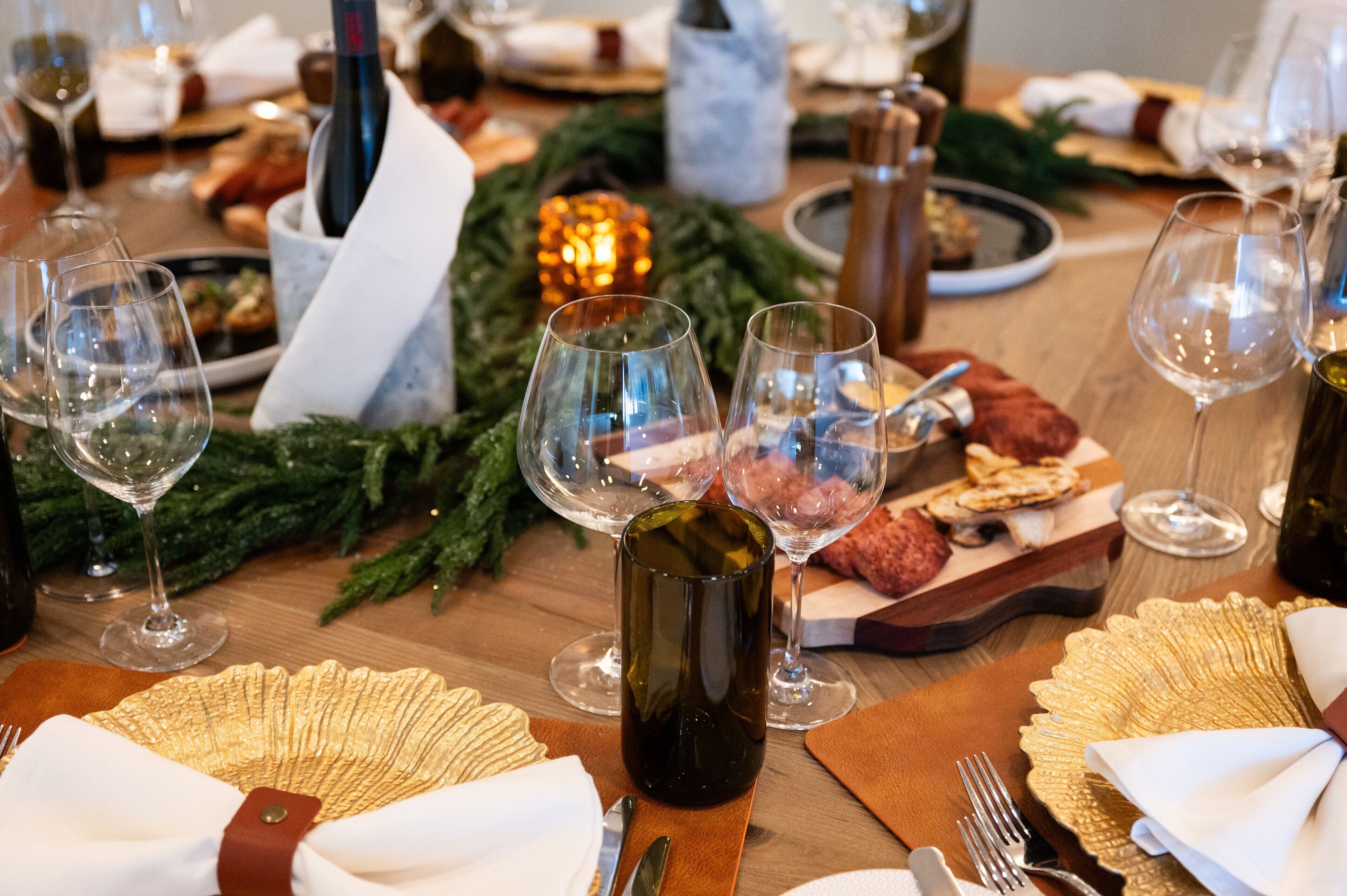 A dining table set with wine glasses, plates, and cutlery, surrounded by greenery and candles.