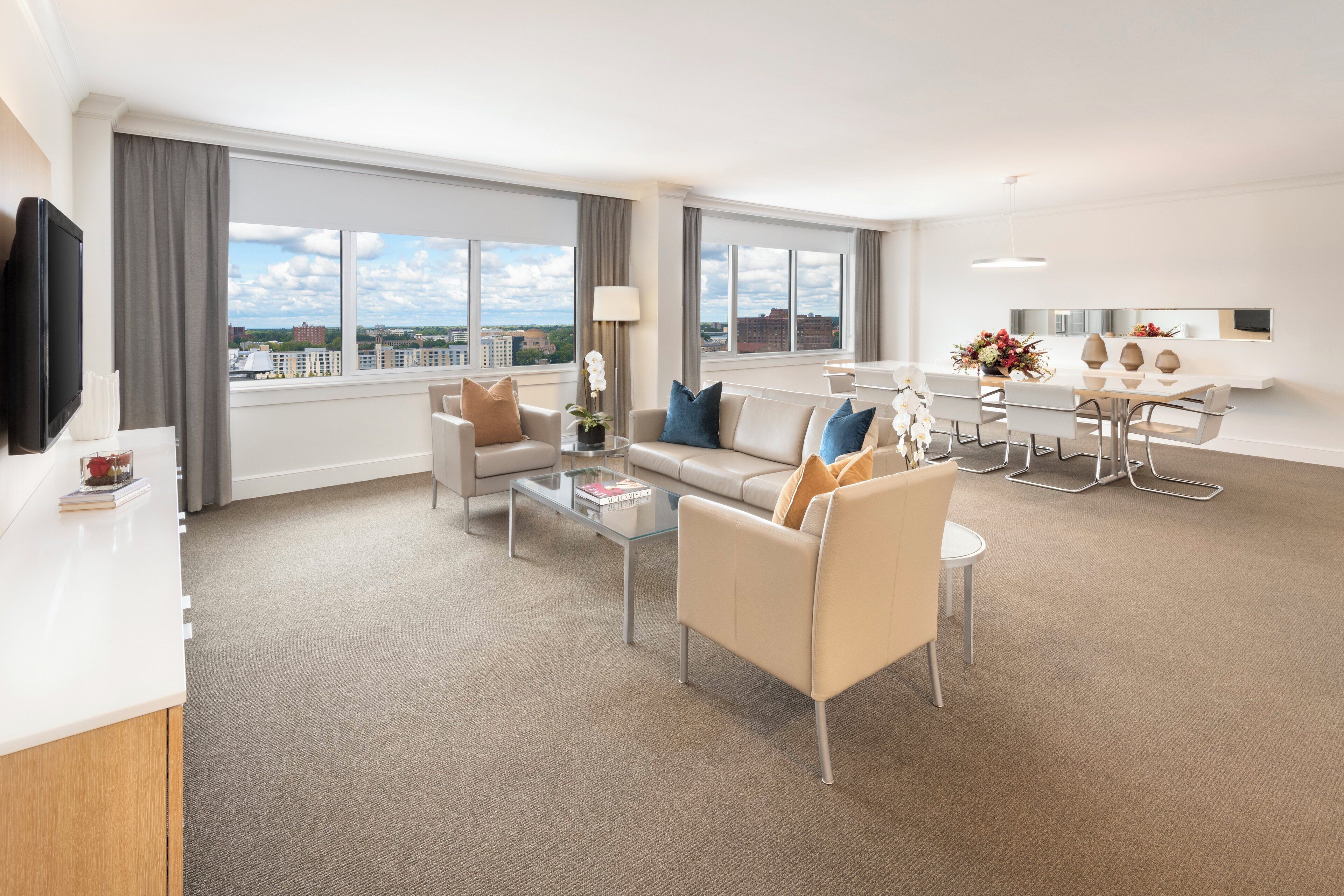 A modern hotel room with a living area, dining space, and large windows offering city views.