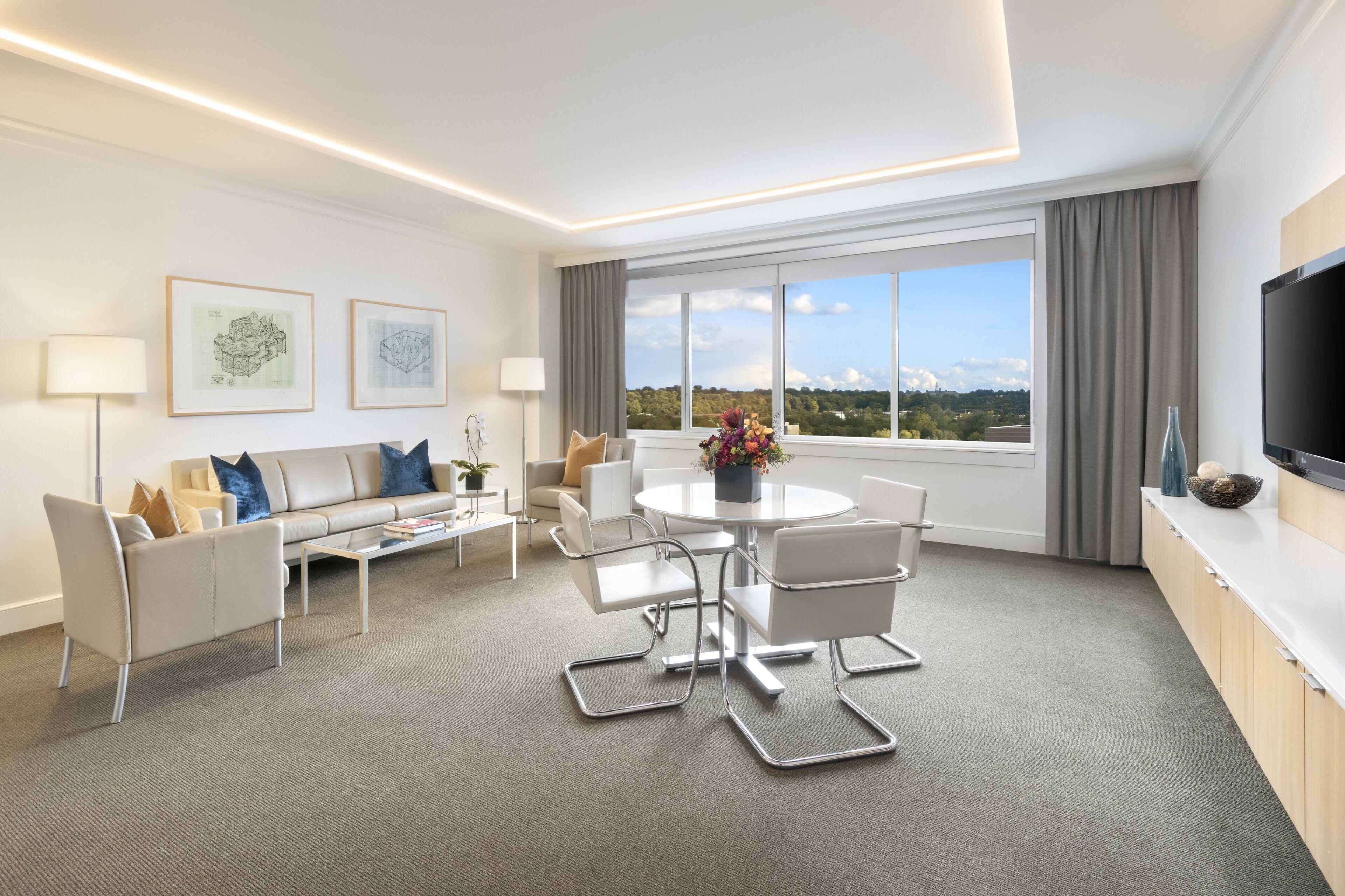 Et moderne hotelværelse med sofa, spisebord, stole, et fjernsyn og et stort vindue med udsigt.