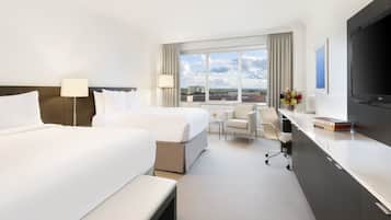 A hotel room with two beds, a desk, a chair, a TV, and a window with a view of the city.