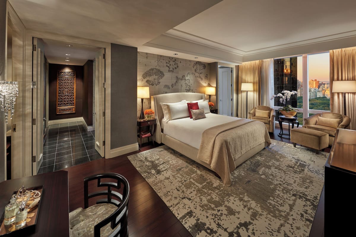 Een ruime hotelkamer met een groot bed, twee nachtkastjes met lampen, een zithoek met een bank en fauteuils, een eettafel met stoelen en een uitzicht op de stad door grote ramen.